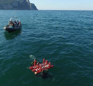 Small red wave device being set up by divers in the ocean, a support vessel drifts nearby