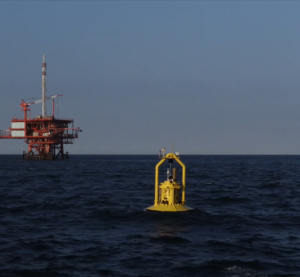 Yellow PowerBuoy floats on the open ocean with an offshore oil rig in the background