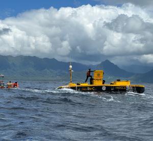 big white clouds hang in the distance over the island of Hawaii, a man climbs onto the hatch of a WEC floating offshore in the blue ocean